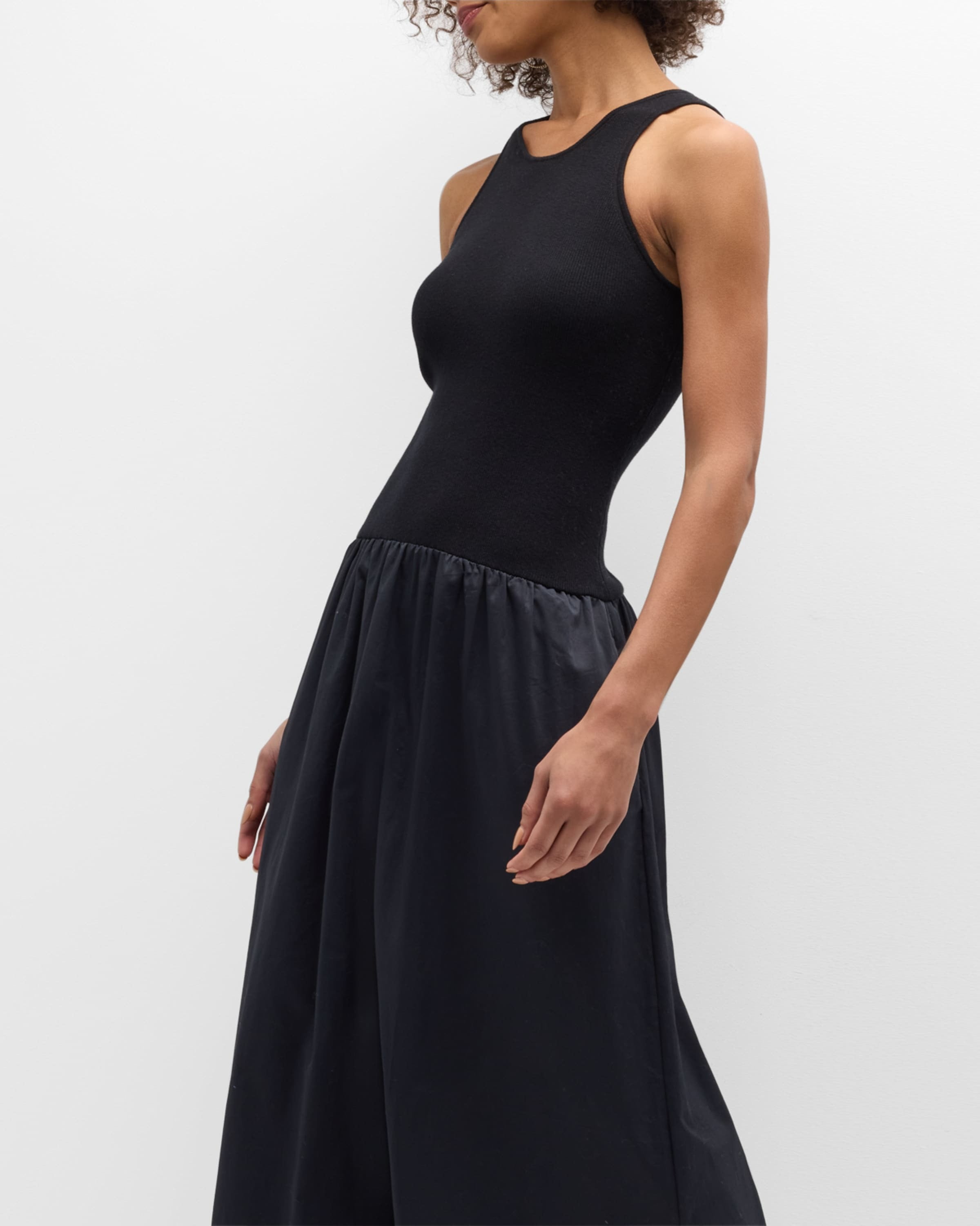 A person is wearing the Naadam Mixed Media Midi Dress in black from Naadam, standing against a plain white background. The dress is fitted at the top with a sleeveless design and features a contrasting, shirred skirt that flows out at the waist. The subject's head is turned to the side, revealing curly hair. The pose is casual and relaxed.