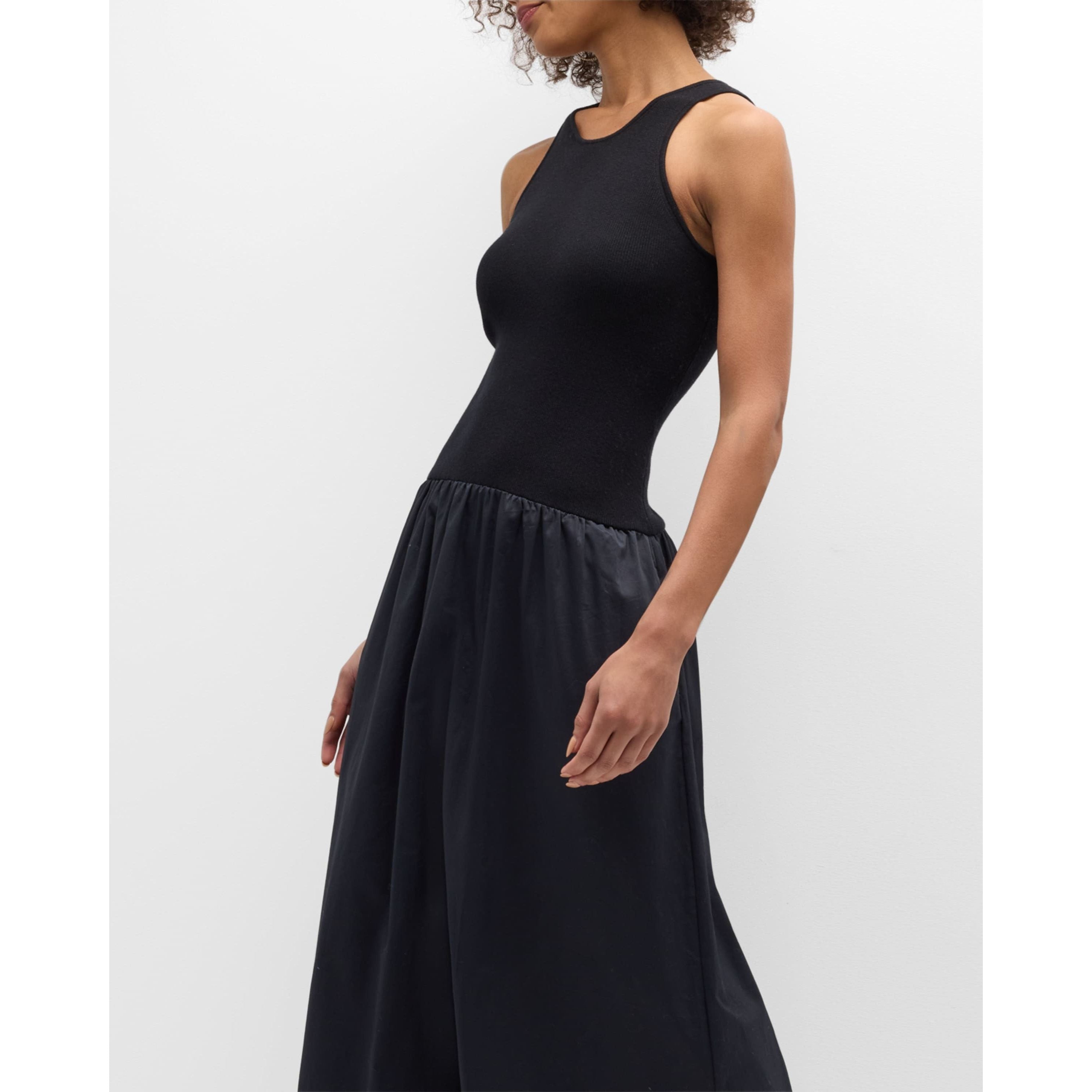 A person is wearing the Naadam Mixed Media Midi Dress in black from Naadam, standing against a plain white background. The dress is fitted at the top with a sleeveless design and features a contrasting, shirred skirt that flows out at the waist. The subject's head is turned to the side, revealing curly hair. The pose is casual and relaxed.