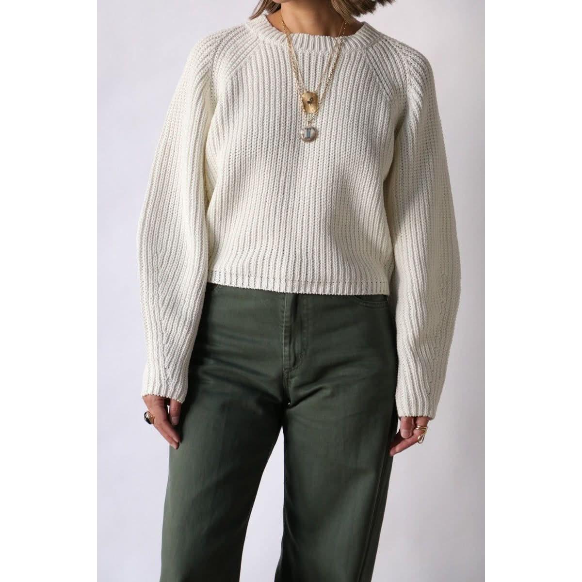 The individual is wearing the Rachel Comey Salla Sweater, a cream-colored chunky knit cotton piece with balloon sleeves, paired with dark green pants. Multiple necklaces elegantly enhance the cropped boxy fit of their outfit. The image captures their stylish ensemble by cropping just above the head.