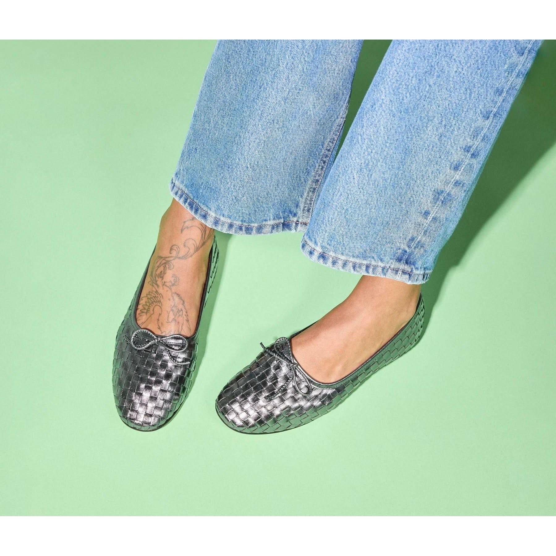 An individual is wearing the Freda Salvador Roma Round Toe Ballet Flats in gunmetal, made from premium Spanish leather, paired with light blue jeans, standing against a light green backdrop. A floral tattoo is visible above the shoe on their left foot.