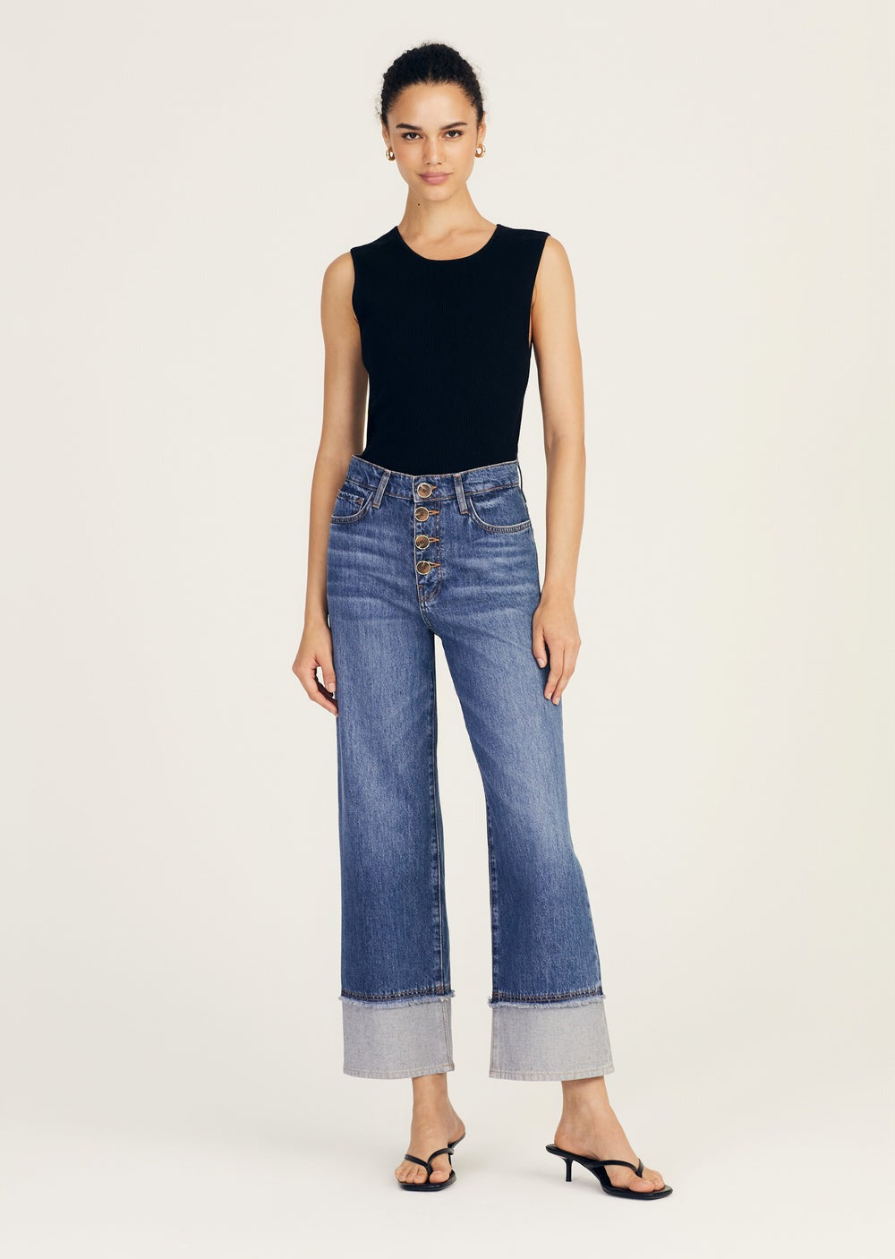 A woman stands confidently against a light background, wearing a sleeveless black top, Derek Lam 10 Crosby Simona Cuffed Straight Leg high-waisted blue jeans with a 5 pocket construction and wide cuffs at the bottom, and black heeled sandals. She has her hair tied back and is staring directly at the camera.