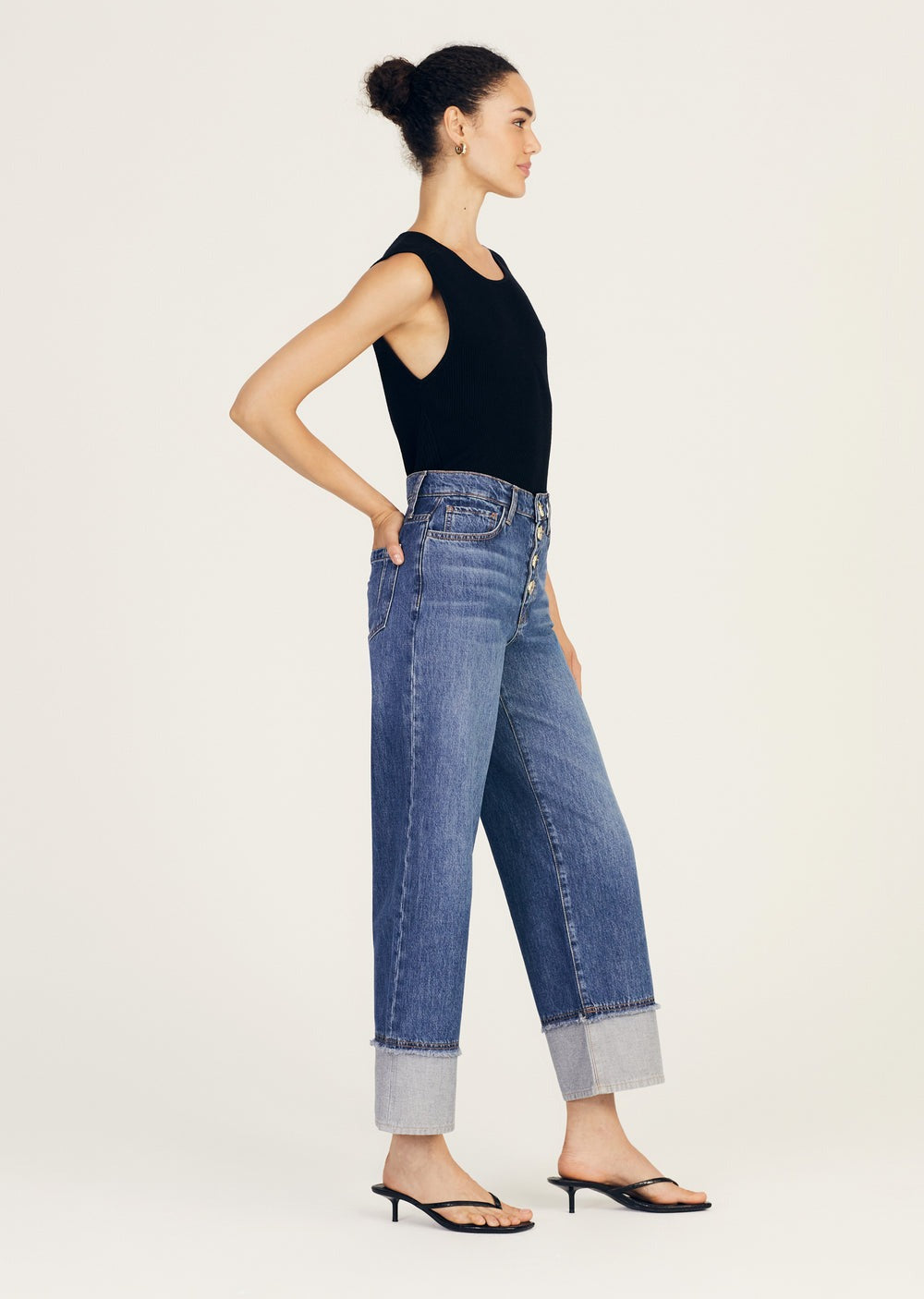 A woman stands in profile against a plain background. She is wearing a sleeveless black top, Derek Lam 10 Crosby Simona Cuffed Straight Leg high-waisted blue jeans with large cuffs at the bottom, and black strappy sandals with a small heel. The rigid denim's 5 pocket construction adds a classic touch to her modern, pulled-together look.