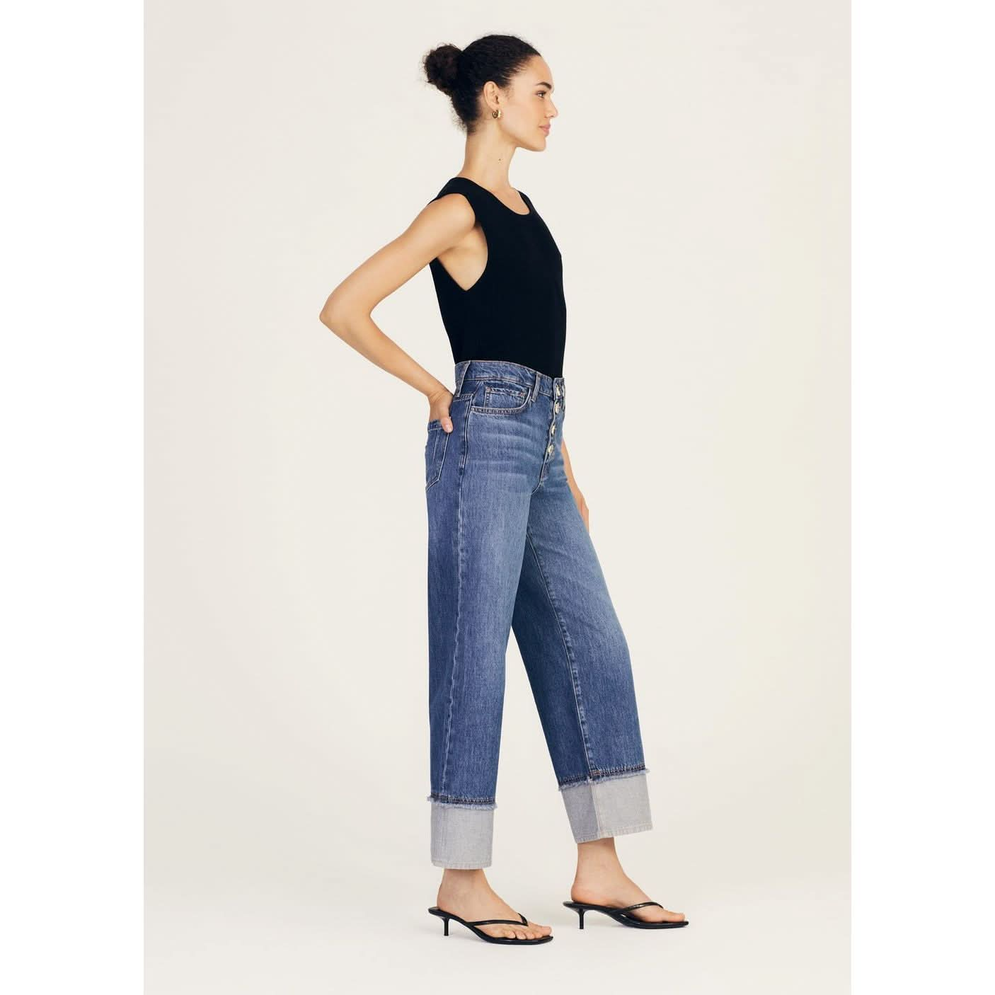 A woman stands in profile against a plain background. She is wearing a sleeveless black top, Derek Lam 10 Crosby Simona Cuffed Straight Leg high-waisted blue jeans with large cuffs at the bottom, and black strappy sandals with a small heel. The rigid denim's 5 pocket construction adds a classic touch to her modern, pulled-together look.