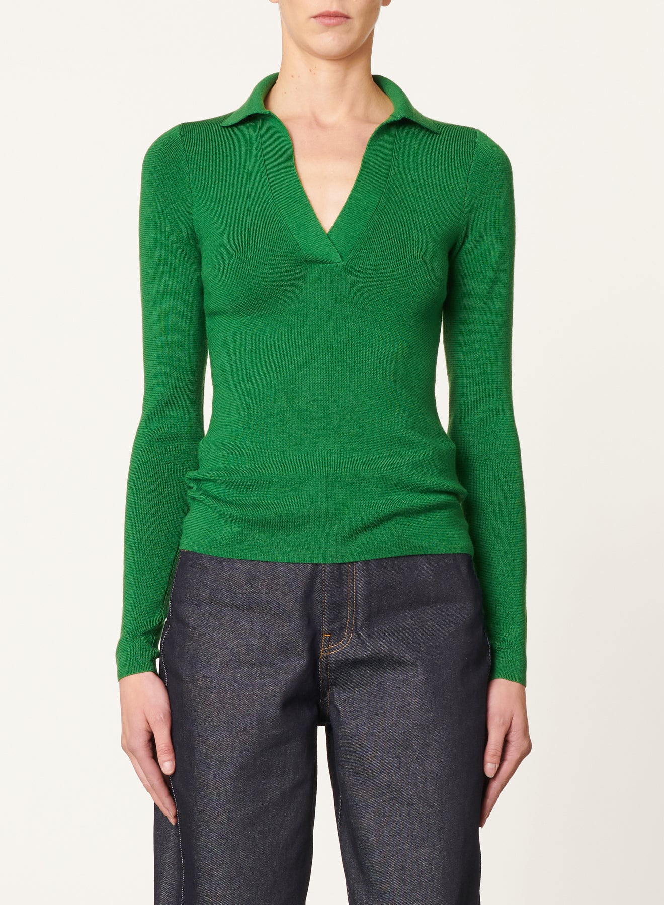 A person is wearing the Vanessa Bruno Dorita Top—a bright green, long-sleeve sweater made of soft wool featuring a collar and a deep V-neckline. They are also dressed in dark blue denim jeans, standing against a plain, light-colored background.