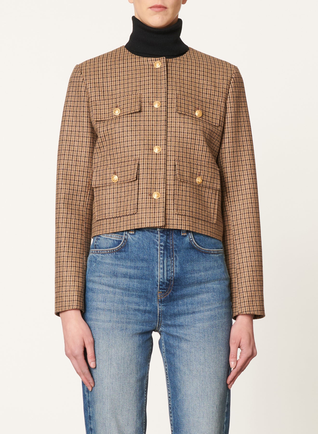 A person is wearing the Vanessa Bruno Deana Jacket, a brown checkered blazer with gold buttons and front pockets, over a black turtleneck. The 100% viscose jacket is paired with blue jeans. The background is plain and off-white.