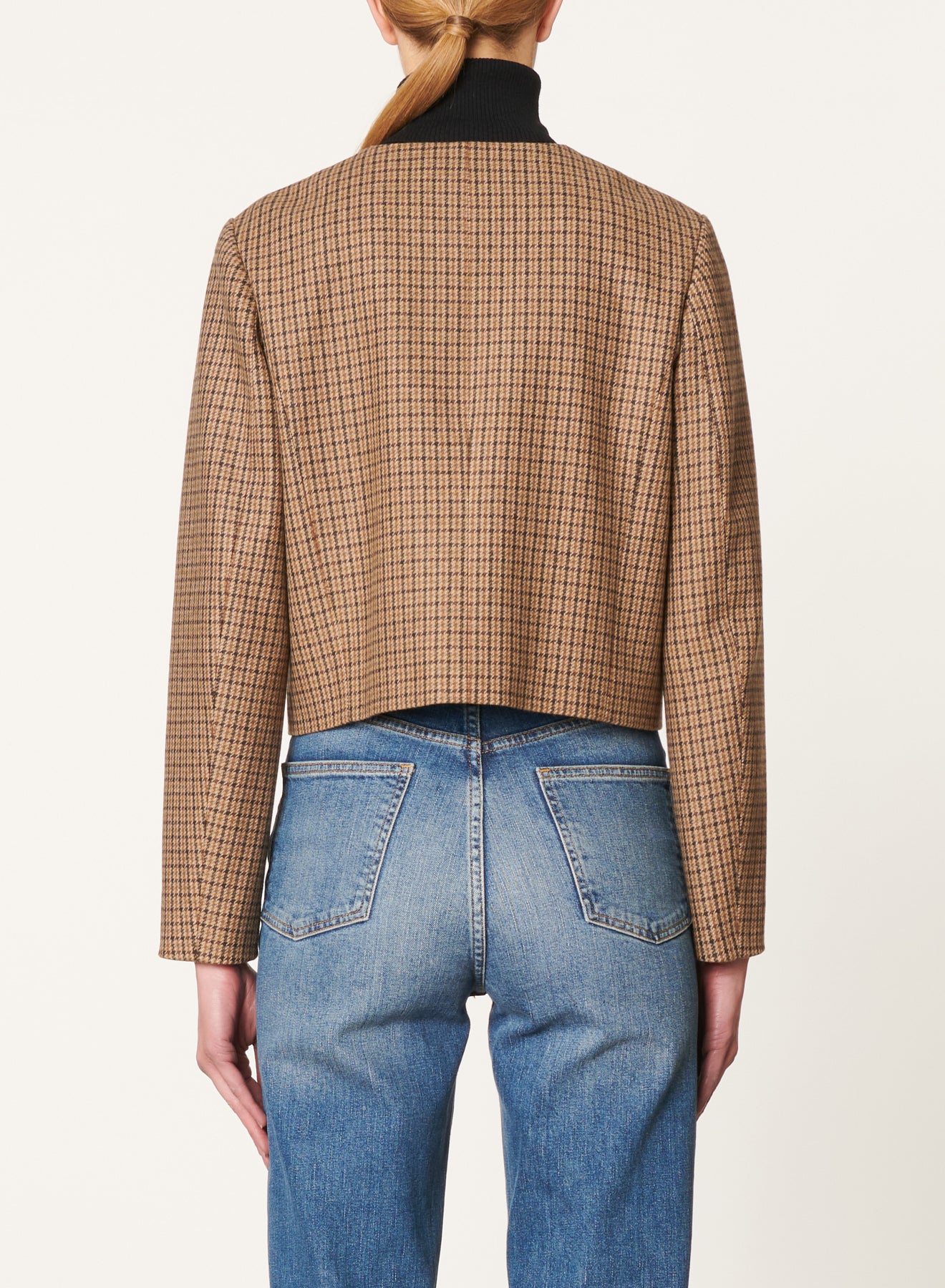 A person with long hair is shown from the back, wearing the Vanessa Bruno Deana Jacket by Vanessa Bruno. Crafted from 100% viscose, this brown plaid jacket features long sleeves and a cropped length that extends just above the waistline of their blue jeans. The background is plain and light-colored.