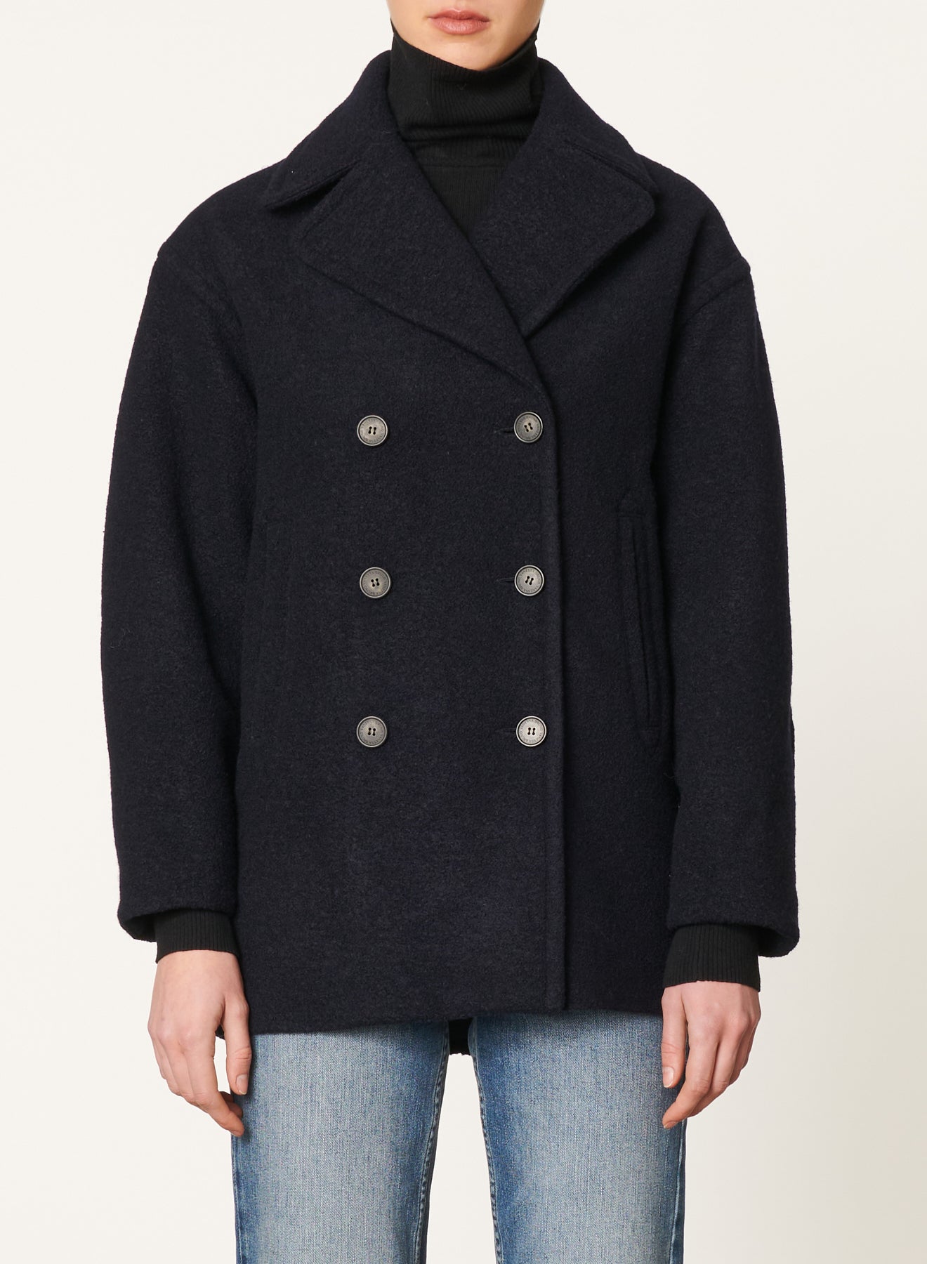 Individual wearing the Vanessa Bruno Berlin Jacket Marine, a dark navy double-breasted peacoat with large buttons, paired with a black turtleneck sweater made from soft 100% viscose. They are also dressed in light blue jeans against an off-white background.