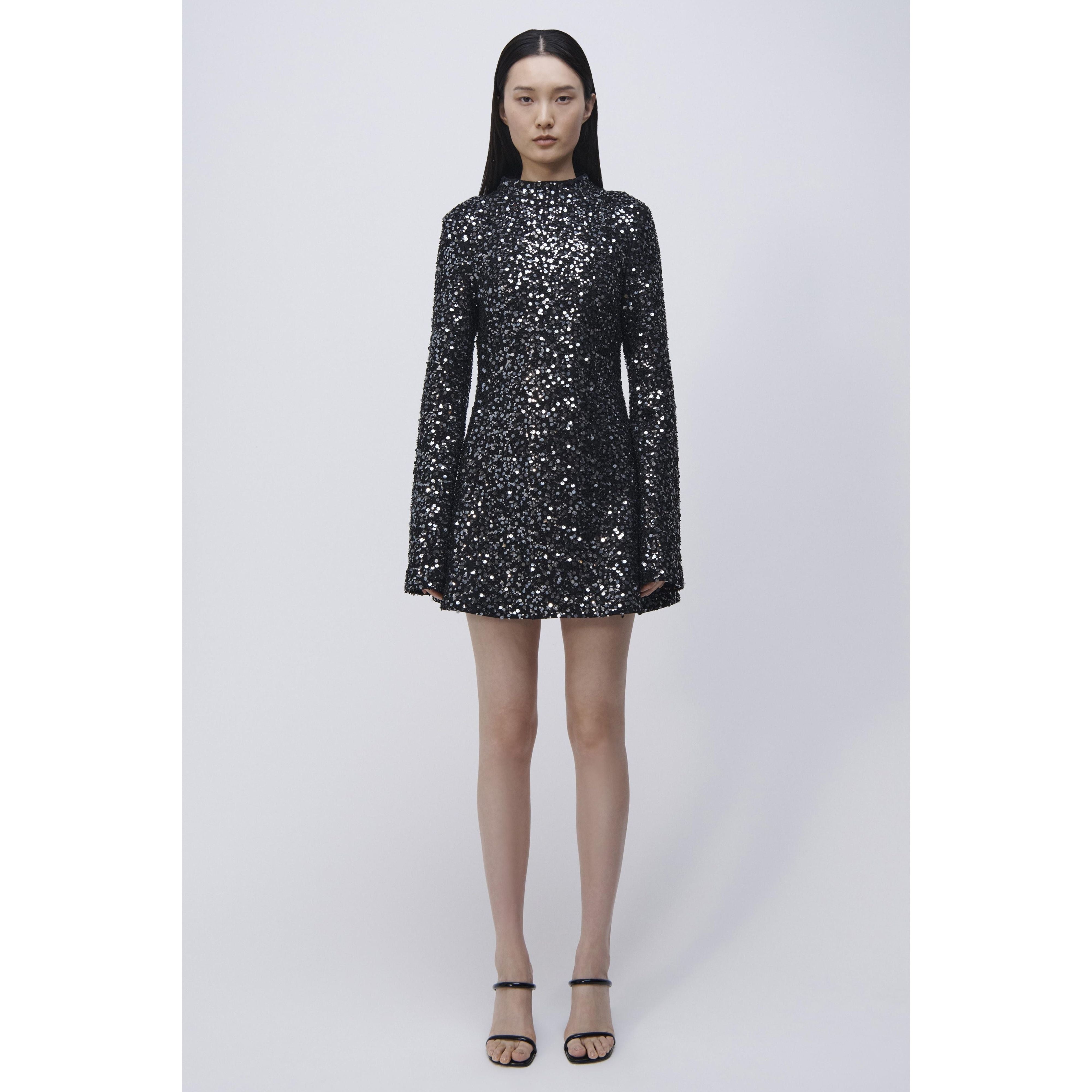 A woman stands against a plain background in a stylish Simkhai Safia Mini Dress. Her long, dark hair cascades straight down, complementing the elegance of her open-toed black high heels. With a neutral expression, she exudes confidence and grace.