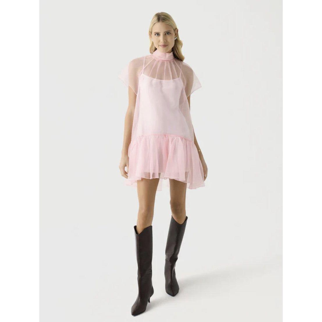 A person is dressed in a Simkhai Beaux Tiered Mini Dress, showcasing a short, flowing pink style with sheer sleeves and a ruffled hem. Their long hair complements the knee-high black boots as they stand against a plain light background.