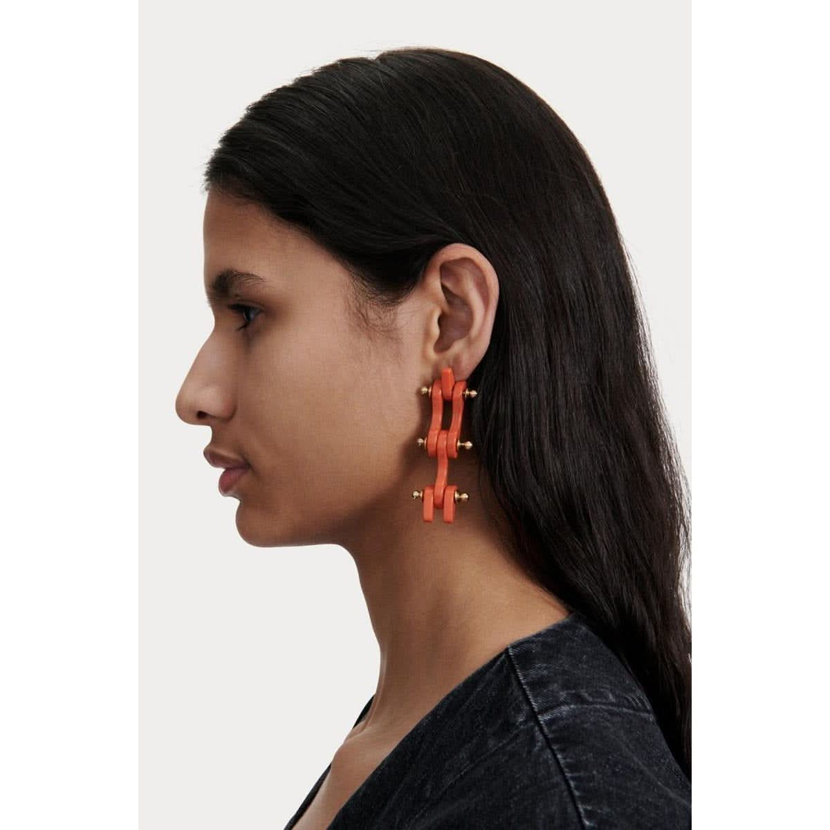 A woman with long black hair is wearing the Rachel Comey Bedford Earrings in Coral, characterized by their chunky, geometric acrylic design. She is shown in profile against a plain white background, dressed in a dark top. The earrings feature surgical steel clips for added comfort.