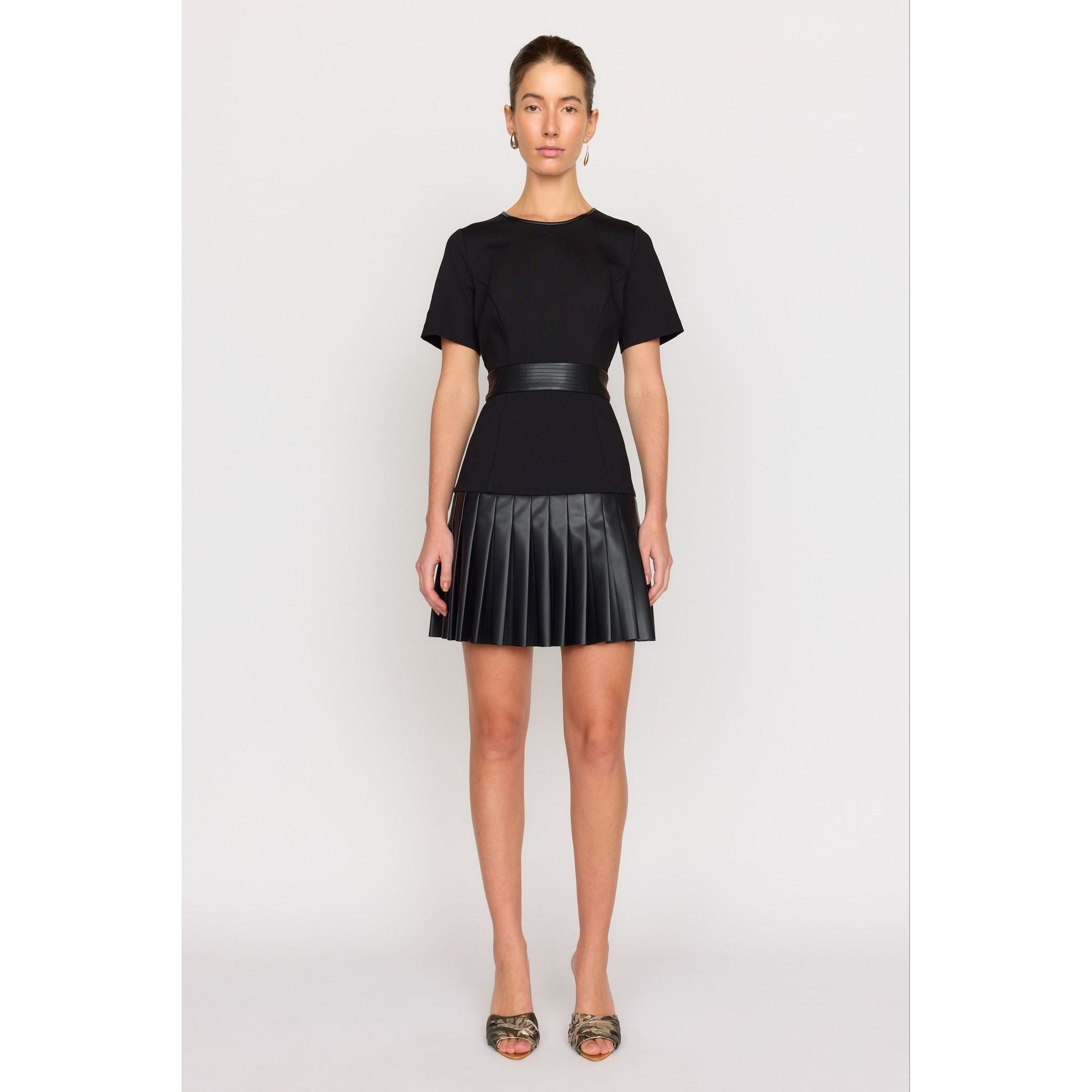 A person stands against a plain background wearing the Christy Lynn Steph Dress, featuring a black short-sleeve top and pleated skirt. Their hair is pulled back, adorned with hoop earrings, and they complete the look with strappy shoes.