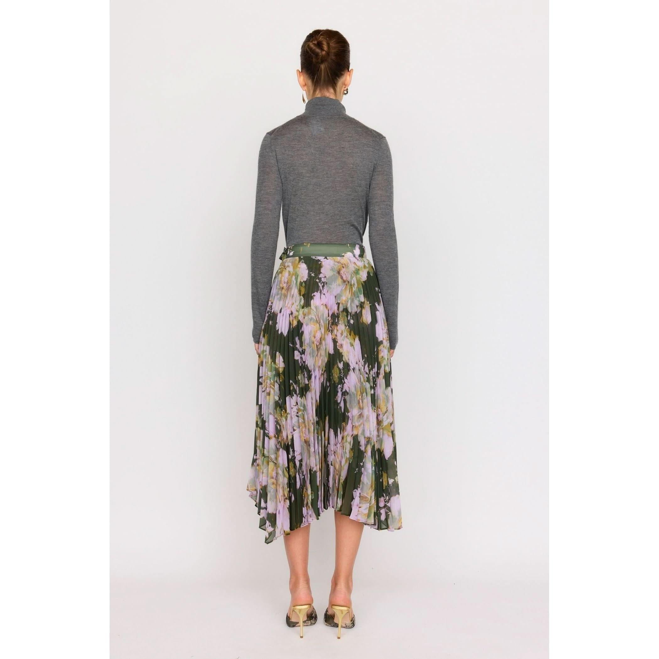 A person stands with their back turned, wearing a long-sleeve gray turtleneck and the Christy Lynn Gwen Skirt in Midnight Garden. The pleated skirt features floral patterns with vibrant green and purple hues, while the fitted waist enhances their silhouette. Their hair is styled in a bun, complemented by beige high-heeled shoes.