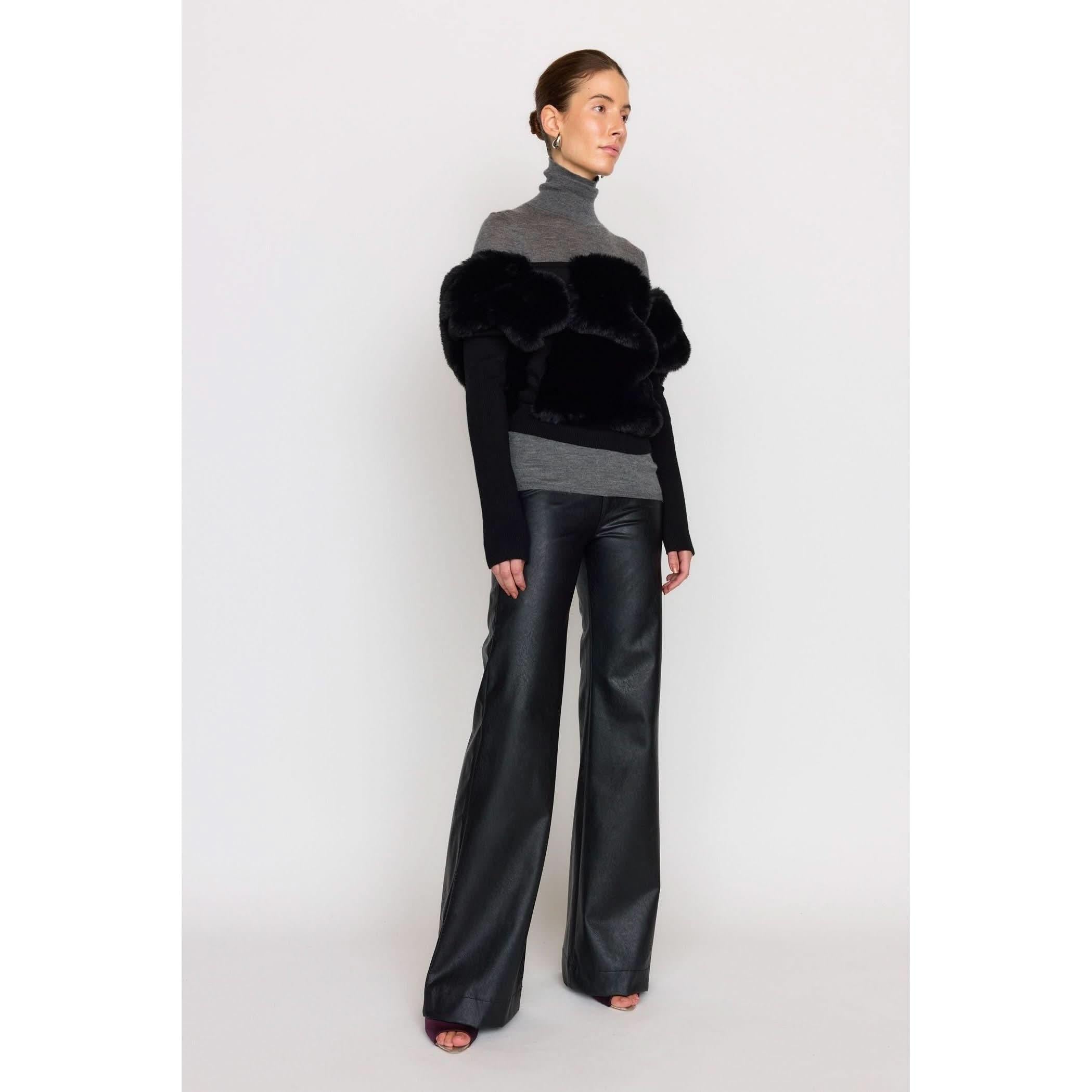 A person stands against a plain background, wearing the stylish Christy Lynn Criselle Knit in black with black fur detail and long sleeves. They also pair this with black wide-leg pants made from vegan leather, creating a modern and chic look.