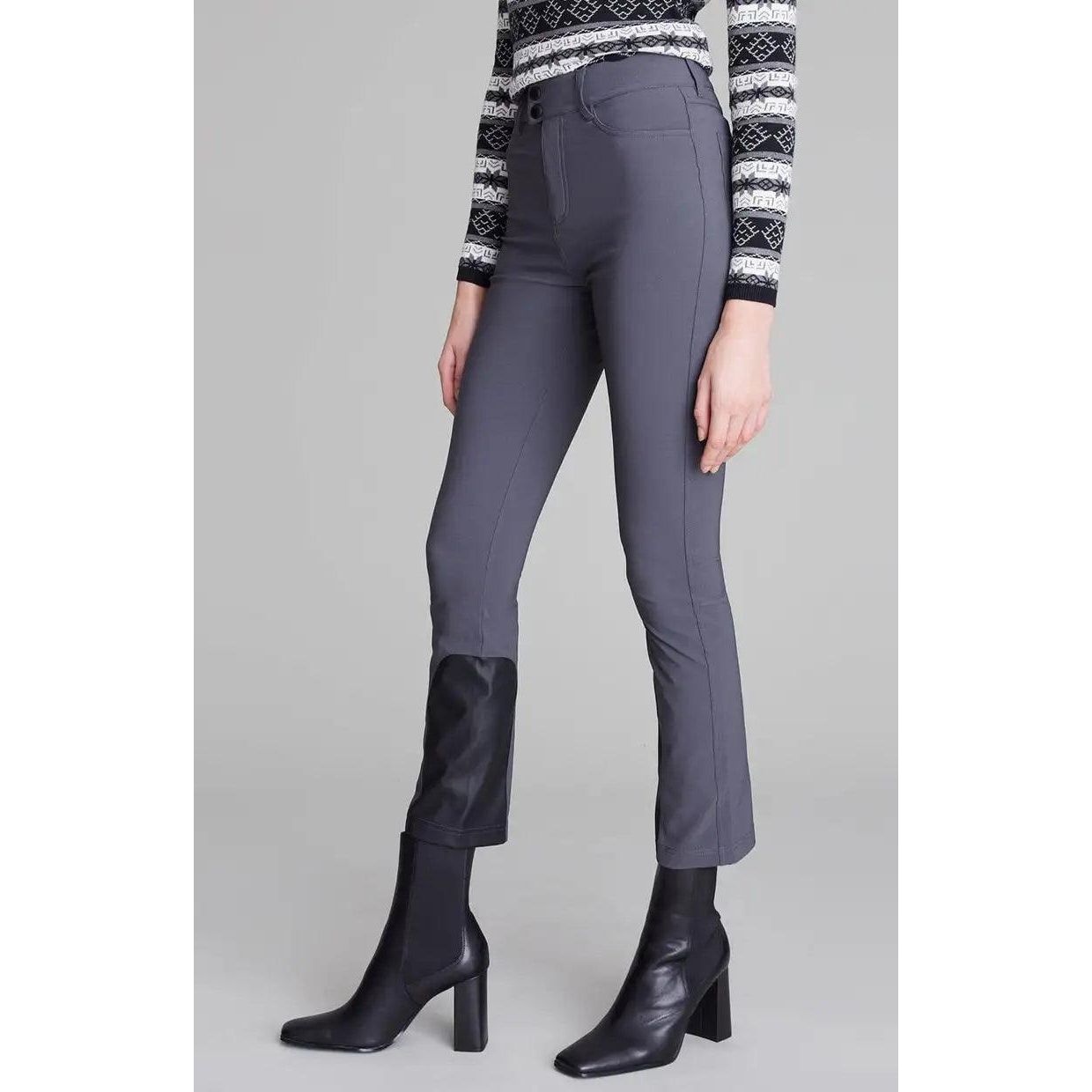 A person is wearing a patterned long-sleeve shirt paired with the Alp N Rock Naomi Straight Leg Pant in Charcoal, which features black vegan leather patches on the lower leg area. They are also sporting black ankle-high boots with block heels, against a plain gray background.