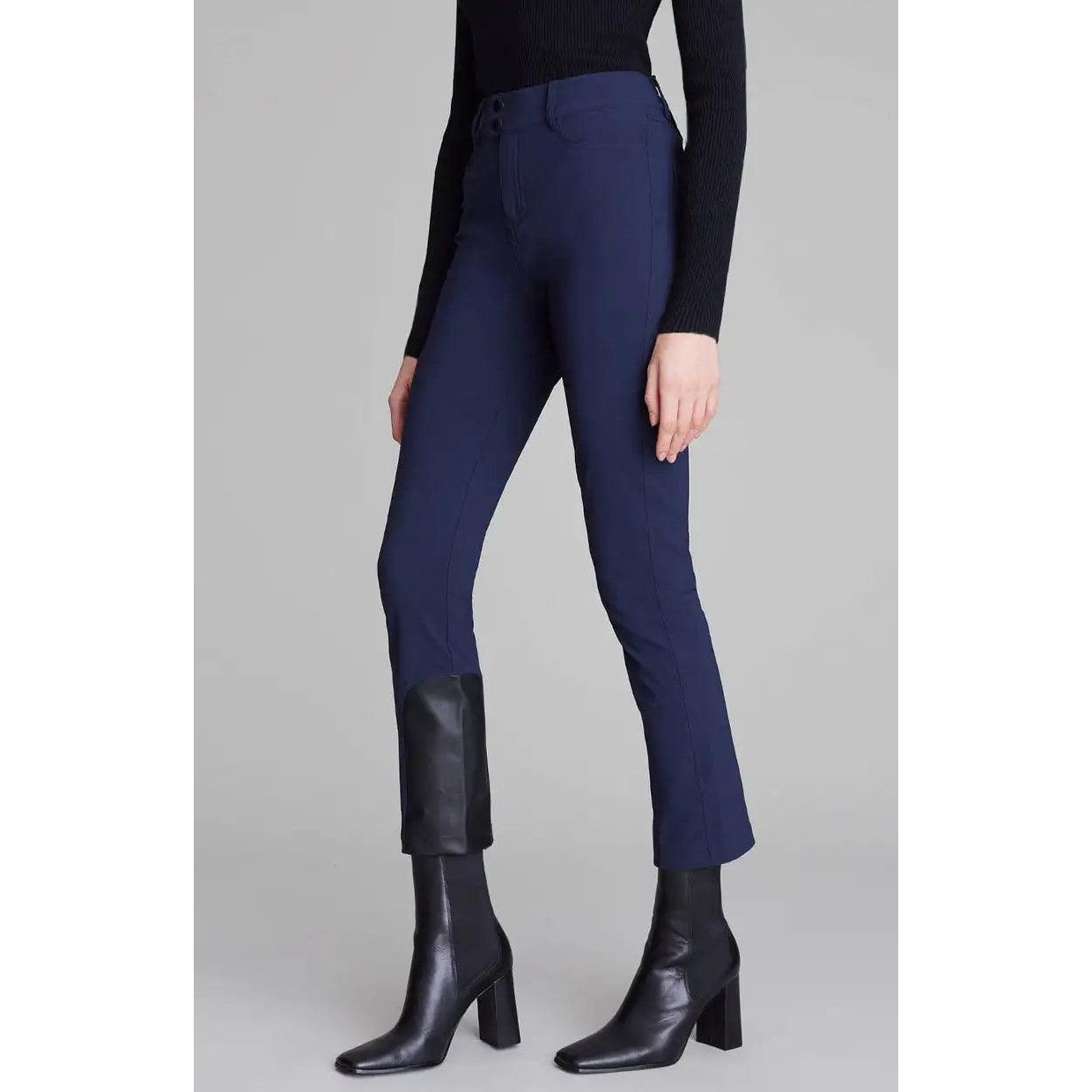 A person is wearing the Alp N Rock Naomi Straight Leg Pant in Dark Sapphire with a black sweater tucked in. The pants, showcasing a slim straight-leg silhouette and split hems at the ankles, reveal black knee-high boots. Subtle vegan leather accents add a touch of sophistication as the person stands against a plain gray background.