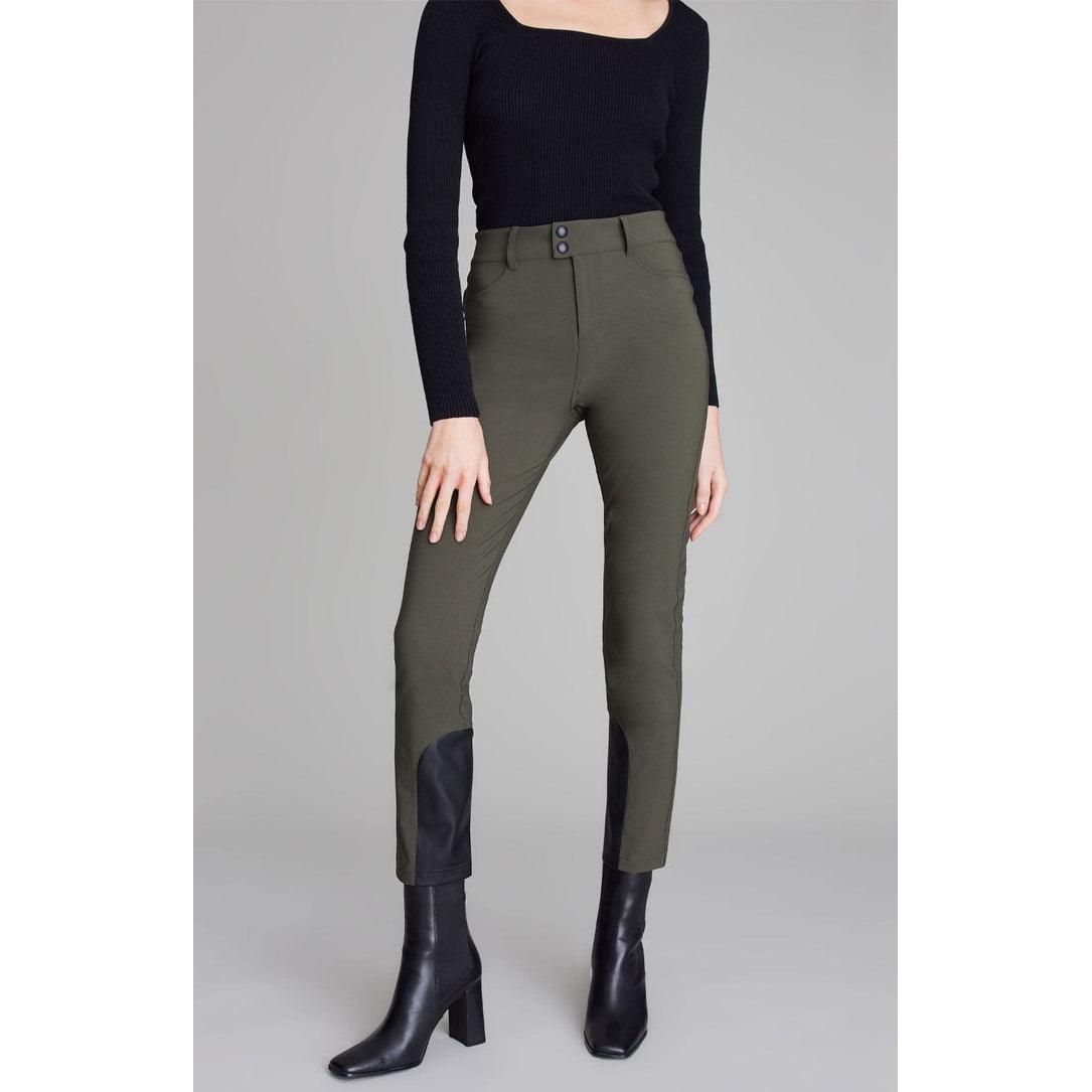 A person is wearing the Alp N Rock Naomi Straight Leg Pant in olive, which features high-waisted slim straight legs with black knee patches and vegan leather accents, paired with a long-sleeved black top. They are also wearing black heeled ankle boots. The background is plain gray. The person's face is not visible in the image.