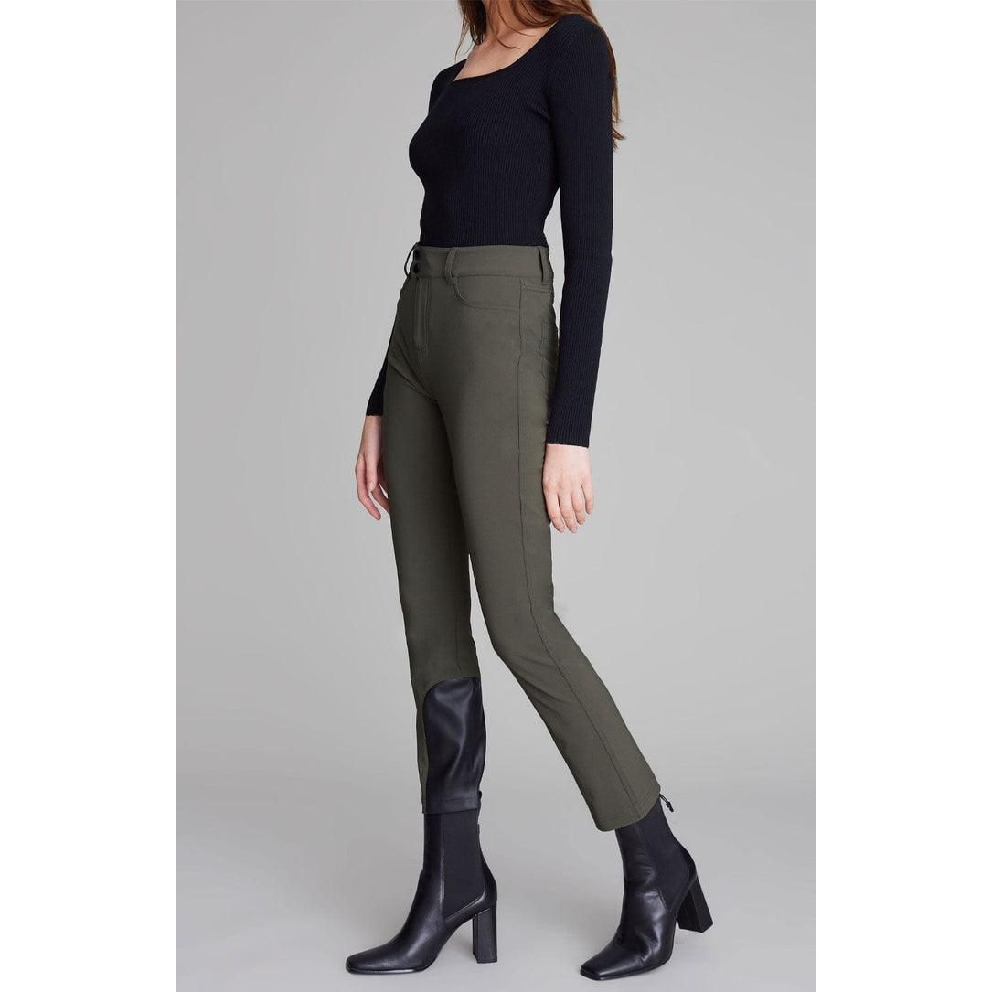 A person stands in a relaxed pose wearing a black long-sleeved top with vegan leather accents, Alp N Rock Naomi Straight Leg Pant in Olive featuring 4-way stretch technology, and black ankle boots with block heels. The background is plain and gray.